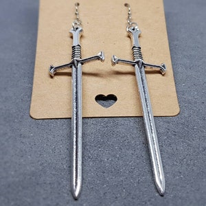 Peut inclure: Une paire de boucles d'oreilles en forme d'&eacute;p&eacute;e argent&eacute;es avec un design d&eacute;taill&eacute;. Les boucles d'oreilles sont suspendues &agrave; une cha&icirc;ne argent&eacute;e.