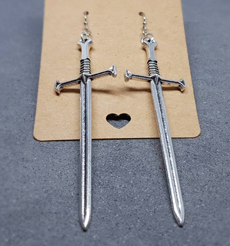 Peut inclure: Une paire de boucles d'oreilles en forme d'&eacute;p&eacute;e argent&eacute;es avec un design d&eacute;taill&eacute;. Les boucles d'oreilles sont suspendues &agrave; une cha&icirc;ne argent&eacute;e.