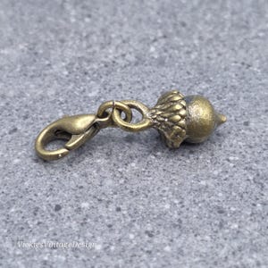 Può includere: Un ciondolo in metallo color bronzo a forma di ghianda con una cupola testurizzata e una parte inferiore liscia, fissato a una chiusura a moschettone tramite un piccolo anellino. Il ciondolo ha una finitura metallica ed è posizionato orizzontalmente.