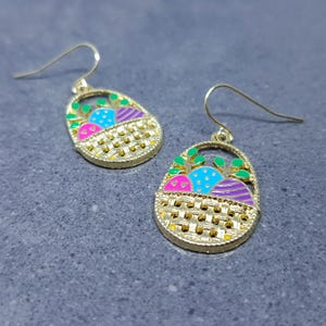 Puede incluir: Pendientes colgantes de tono dorado con un diseño de cesta. La cesta está llena de huevos de Pascua coloridos y hojas verdes.