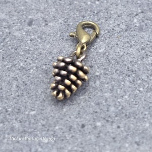 May include: A bronze-toned metal charm shaped like a pinecone, featuring a textured surface with individual scales. The charm is attached to a small lobster claw clasp, allowing it to be easily clipped onto jewellery, keychains, or other accessories.