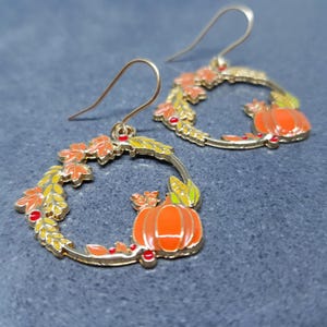 May include: Gold-coloured metal earrings with a circular design. They feature orange pumpkins, autumn leaves, and red berries. The earrings have a hook closure. The earrings are a fall-themed accessory, set against a grey background.
