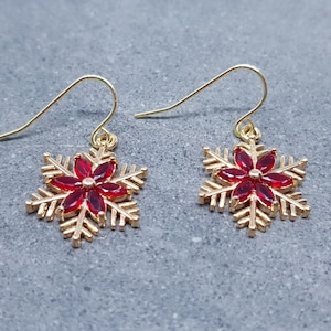May include: Gold-coloured snowflake earrings with red gemstone detailing. The earrings feature a hook fastening and are set against a grey background. The snowflake design is a festive holiday theme.