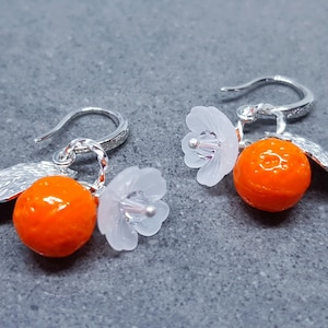 May include: A pair of silver earrings with orange and white charms. The orange charms are shaped like oranges and the white charms are shaped like flowers.