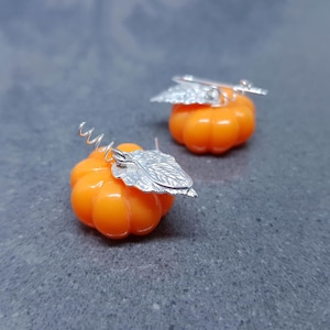 Op de afbeelding: Een paar oranje pompoen oorbellen met zilverkleurige blad- en krulaccenten. De oorbellen zijn ontworpen om op miniatuur pompoenen te lijken, perfect voor de herfst of Halloween.