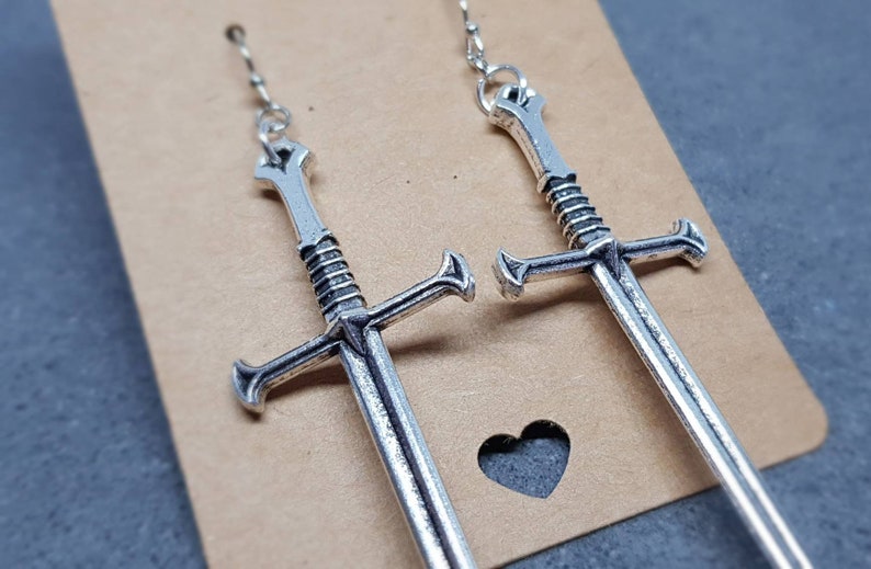 Peut inclure: Une paire de boucles d'oreilles en forme d'&eacute;p&eacute;e argent&eacute;es avec un design d&eacute;taill&eacute;. Les &eacute;p&eacute;es sont suspendues &agrave; une cha&icirc;ne et mesurent environ 7,5 cm de long.