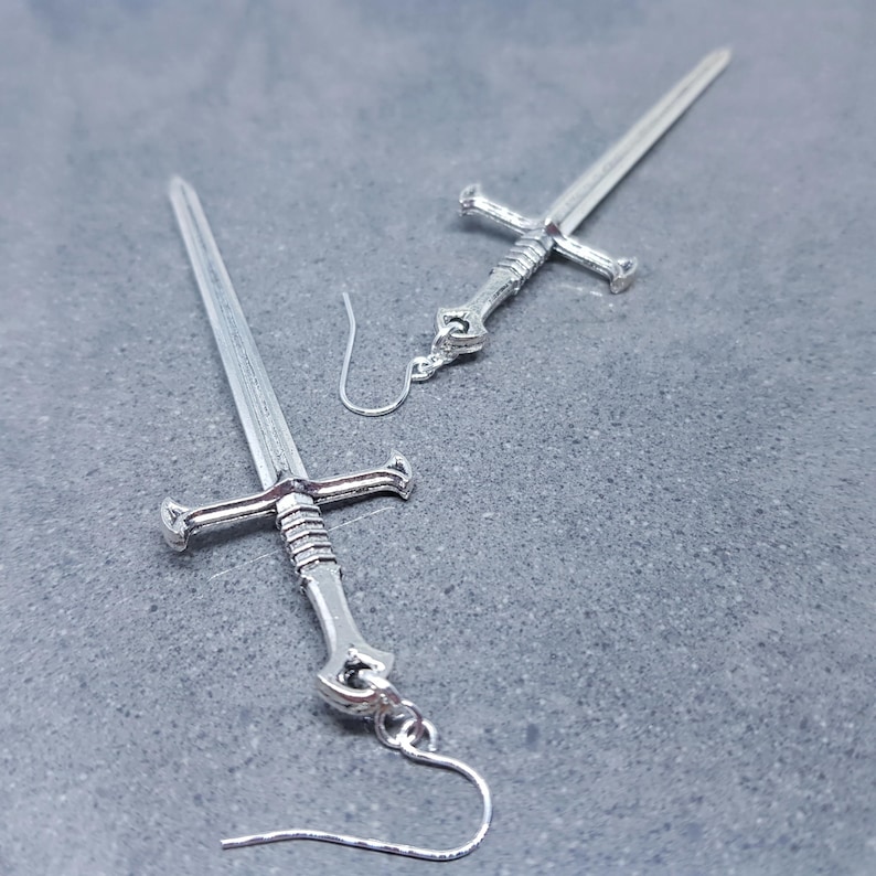 K&ouml;nnte beinhalten: Ein Paar silberne Schwertohrringe. Die Schwerter haben ein detailliertes Design und h&auml;ngen an silbernen Haken.