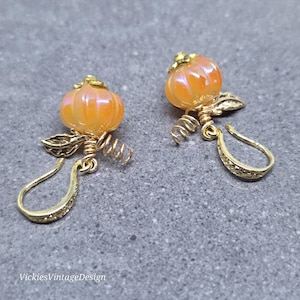 May include: A pair of earrings featuring iridescent orange pumpkin-shaped beads with gold-tone metal leaves and coiled wire accents. The earrings have gold-tone hooks with a textured finish. The text VickiesVintageDesign is visible in the corner.