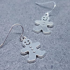 May include: A pair of silver dangle earrings featuring gingerbread man charms. The gingerbread men have a smiling face and button details.