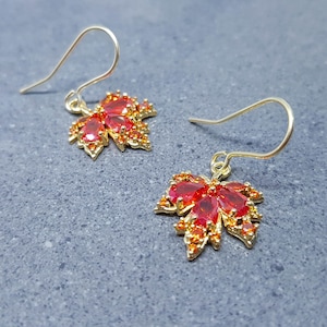 Op de afbeelding: Een paar goudkleurige oorbellen in de vorm van een blad. Elke oorbel heeft een cluster van rode en oranje edelstenen, met een haakje voor doorboorde oren. De oorbellen staan tegen een grijze achtergrond.