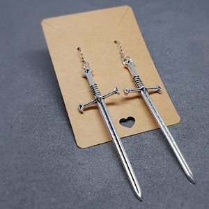 Peut inclure: Une paire de boucles d'oreilles en forme d'&eacute;p&eacute;e argent&eacute;es suspendues &agrave; une carte marron. Les &eacute;p&eacute;es ont un design d&eacute;taill&eacute; et mesurent environ 7,5 cm de long.