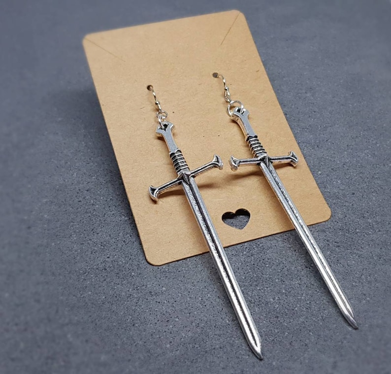 Peut inclure: Une paire de boucles d'oreilles en forme d'&eacute;p&eacute;e argent&eacute;es suspendues &agrave; une carte marron. Les &eacute;p&eacute;es ont un design d&eacute;taill&eacute; et mesurent environ 7,5 cm de long.