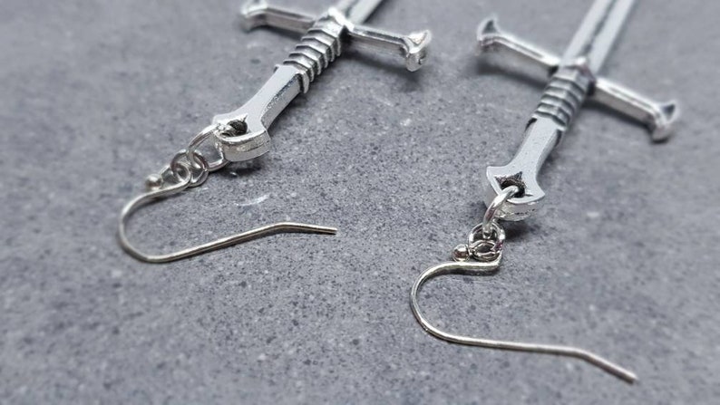 Peut inclure: Une paire de boucles d'oreilles en forme d'&eacute;p&eacute;e argent&eacute;es avec un design d&eacute;taill&eacute;. Les boucles d'oreilles ont une fermeture &agrave; crochet.