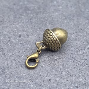 Può includere: Un ciondolo in metallo color ottone antico a forma di ghianda, con una cupola testurizzata e una parte liscia, fissato a un piccolo moschettone. Il testo "VickiesVintageDesign" è visibile nell'angolo in basso a sinistra dell'immagine.
