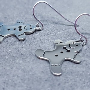 May include: A pair of silver-colored gingerbread man earrings. Each earring features a smiling gingerbread man with small dot and line details. The earrings hang from silver-colored fish hook ear wires. The background is a gray, textured surface.