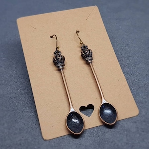 May include: A pair of spoon earrings with a crown design. The spoons are made of a dark metal with a black finish. The crowns are gold coloured.