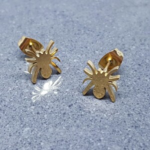 May include: A pair of gold-coloured spider stud earrings. Each earring features a detailed spider silhouette with eight legs and a round body. The earrings have a post and backing for secure wear. The earrings are displayed on a grey surface.