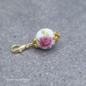 May include: A gold-toned metal charm featuring a round white bead decorated with a pink rose and green leaves. The bead is flanked by gold-toned metal floral caps and attached to a gold-toned lobster clasp. The text VickiesVintageDesign is visible.