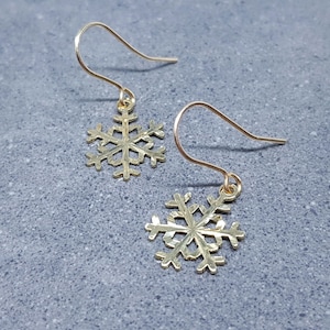 Könnte beinhalten: Ein Paar goldfarbene Schneeflocken-Ohrringe. Jeder Ohrring hat ein filigranes Schneeflocken-Design, das an einem einfachen Haken hängt. Die Ohrringe werden auf einer strukturierten grauen Oberfläche präsentiert.