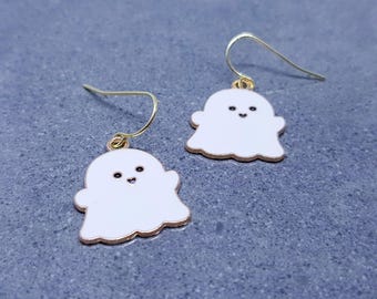 May include: A pair of white ghost-shaped earrings with gold-colored hooks. Each ghost has a gold-colored outline and a smiling face with black eyes and a mouth. The earrings are designed for pierced ears.