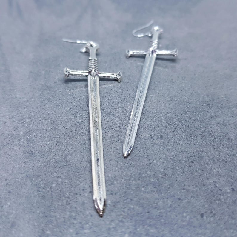 K&ouml;nnte beinhalten: Ein Paar silberne Schwertohrringe. Die Schwerter haben ein detailliertes Design und sind etwa 5 cm lang.