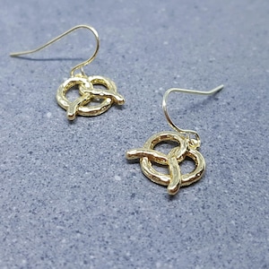 Op de afbeelding: Een paar gouden hangende oorbellen met een pretzel-ontwerp. De oorbellen hebben een textuur en zijn perfect om een ​​vleugje whimsy toe te voegen aan elke outfit.