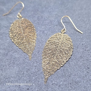 May include: A pair of gold-coloured leaf-shaped earrings. The earrings feature a delicate, openwork design, resembling the veins of a leaf. They have a hook closure. The brand name "VickiesVintageDesign" is visible in the bottom left corner.