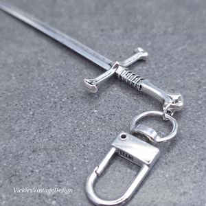 May include: A silver sword-shaped keyring with a detailed hilt and blade. The keyring includes a clasp and ring for attaching to keys or a bag. The text "VickiesVintageDesign" is visible in the lower left corner.
