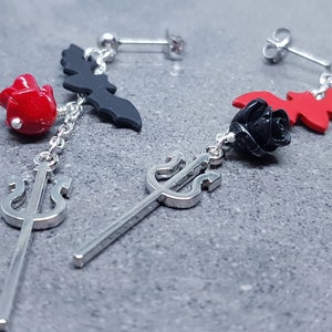 May include: A pair of dangle earrings featuring a black bat, a red rose, and a silver trident charm. The earrings are silver and have a post back.