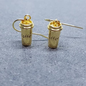 Puede incluir: Un par de pendientes de color dorado en forma de vasos de café con la palabra "coffee" inscrita en la parte delantera.