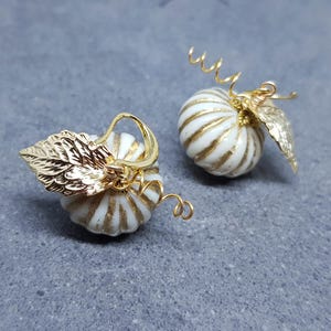 May include: A pair of gold pumpkin earrings with white and gold striped details. The pumpkins have a small gold leaf attached to the stem.