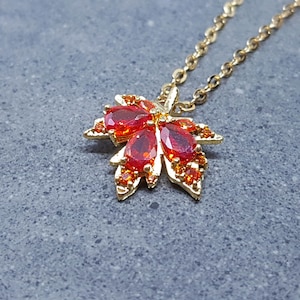 Puede incluir: Un collar dorado con un colgante de hoja de arce. El colgante está adornado con piedras preciosas rojas y naranjas, creando un diseño vibrante. El collar se muestra sobre una superficie gris.