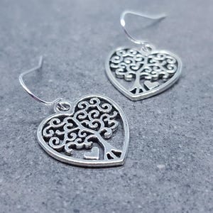 May include: A pair of silver earrings with a heart-shaped design featuring a tree of life inside. The earrings have a delicate, intricate design.