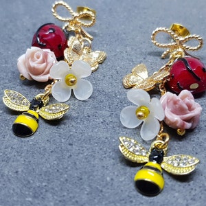 以下が含まれることがあります： 蜂、テントウムシ、花のチャームが付いたゴールドのドロップイヤリング。蜂のチャームは黄色と黒、テントウムシのチャームは黒点のある赤、花のチャームはピンクと白です。
