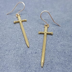 以下が含まれることがあります： 金色の剣のイヤリングのペア。各イヤリングは、十字型の柄と尖った刃が付いた詳細な剣のペンダントが特徴です。イヤリングは、ピアス用のシンプルなフックデザインです。