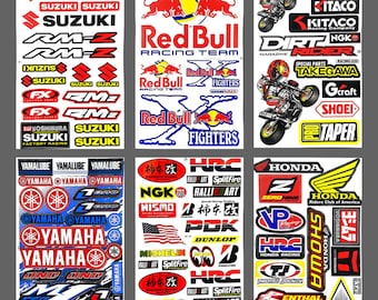 May include: A sheet of colorful motorcycle racing decals featuring logos for brands like Red Bull, Suzuki, Yamaha, Honda, and NGK. The decals are printed on white paper and are ready to be applied to a motorcycle.
