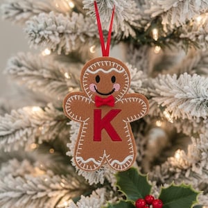 May include: A brown felt gingerbread man ornament featuring white stitched detailing, pink embroidered cheeks, a small red bow tie, and a large red letter K in the center. The ornament hangs from a thin red ribbon loop for display purposes.