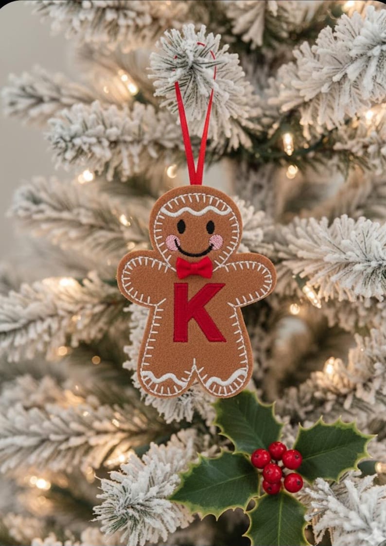 May include: A brown felt gingerbread man ornament featuring white stitched detailing, pink embroidered cheeks, a small red bow tie, and a large red letter K in the center. The ornament hangs from a thin red ribbon loop for display purposes.