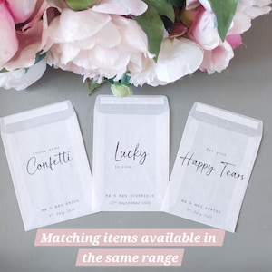 May include: Three translucent white paper envelopes with black printed text. The first reads Throw some Confetti, Mr & Mrs Smith, 9th July 2026. The second reads Lucky in love, Mr & Mrs Schofield, 22nd September 2026. The third reads For your Happy Tears, Mr & Mrs Keates, 7th July 2027.
