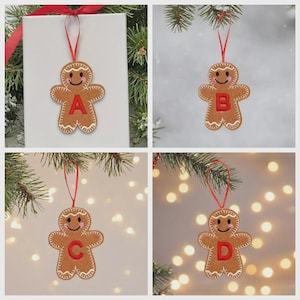 May include: A brown gingerbread man ornament featuring a red embroidered letter on the chest, white stitched icing details, pink cheeks, and a red ribbon loop for hanging. The ornament is shown in four variations labeled with the letters A, B, C, and D.