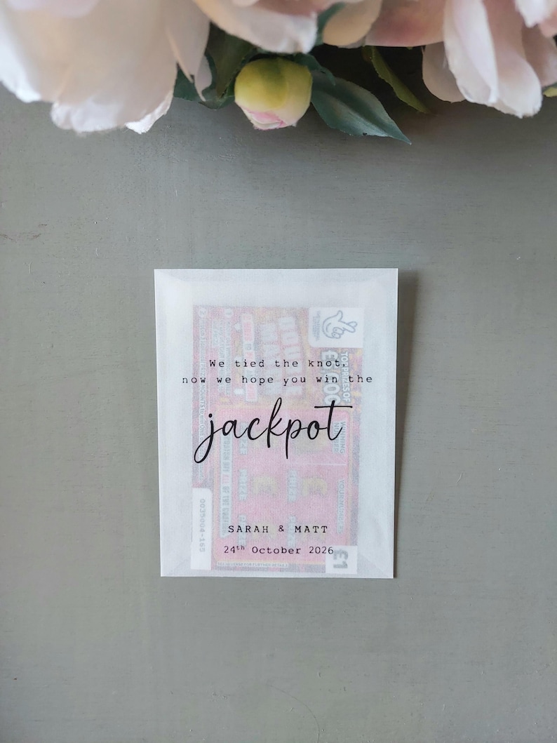 May include: A translucent white paper envelope containing a lottery ticket. The front features black printed text that reads: We tied the knot, now we hope you win the jackpot, Sarah & Matt, 24th October 2026. The envelope is rectangular and flat.