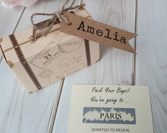 Op de afbeelding: Een klein bruin koffertje in de vorm van een geschenkdoos met een label waarop "Amelia" staat. De doos heeft een vintage reisthema met postzegels en een "Bon Voyage"-label. Een aparte kaart zegt "Pak je tassen! Je gaat naar... PARIJS" met een krasgedeelte om de bestemming te onthullen.