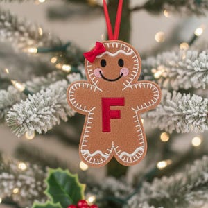 May include: A tan felt gingerbread person ornament featuring a red letter F in the center, a small red bow on the head, white stitched detailing along the edges, and pink cheeks. The ornament hangs from a thin red ribbon loop.