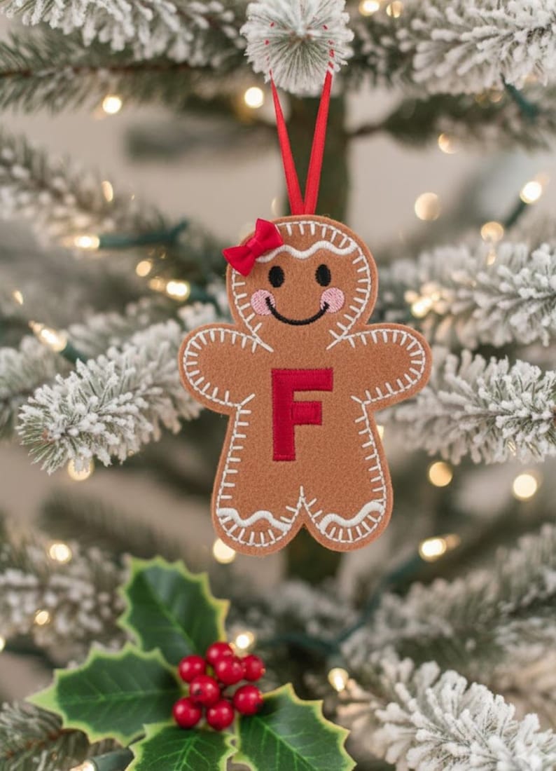 May include: A tan felt gingerbread person ornament featuring a red letter F in the center, a small red bow on the head, white stitched detailing along the edges, and pink cheeks. The ornament hangs from a thin red ribbon loop.