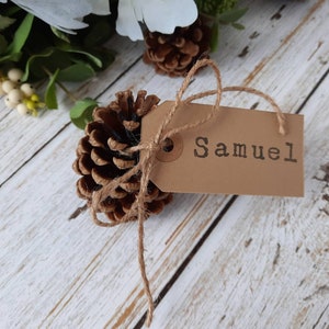 May include: A brown pine cone with a brown paper tag tied to it with twine. The tag has the name "Samuel" written on it.