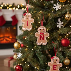 May include: Three felt gingerbread person ornaments hanging on a Christmas tree. Each ornament features white stitched icing details, a red ribbon loop, and a single red letter on the chest spelling out the word INE in a vertical arrangement.