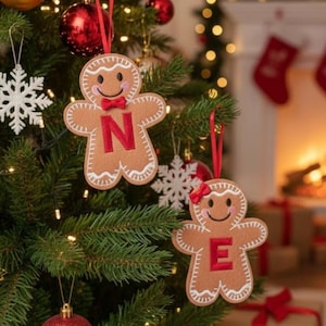 May include: Two tan felt gingerbread person ornaments hanging on a Christmas tree. One ornament features a red letter N and a red bow tie, while the other features a red letter E and a red hair bow. Both have white stitched details and red ribbons.