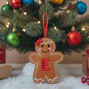 May include: A brown felt gingerbread person ornament featuring a red letter E on the chest, a small red bow on the head, pink cheeks, and white embroidered icing details. The ornament hangs from a red ribbon loop against a Christmas tree.