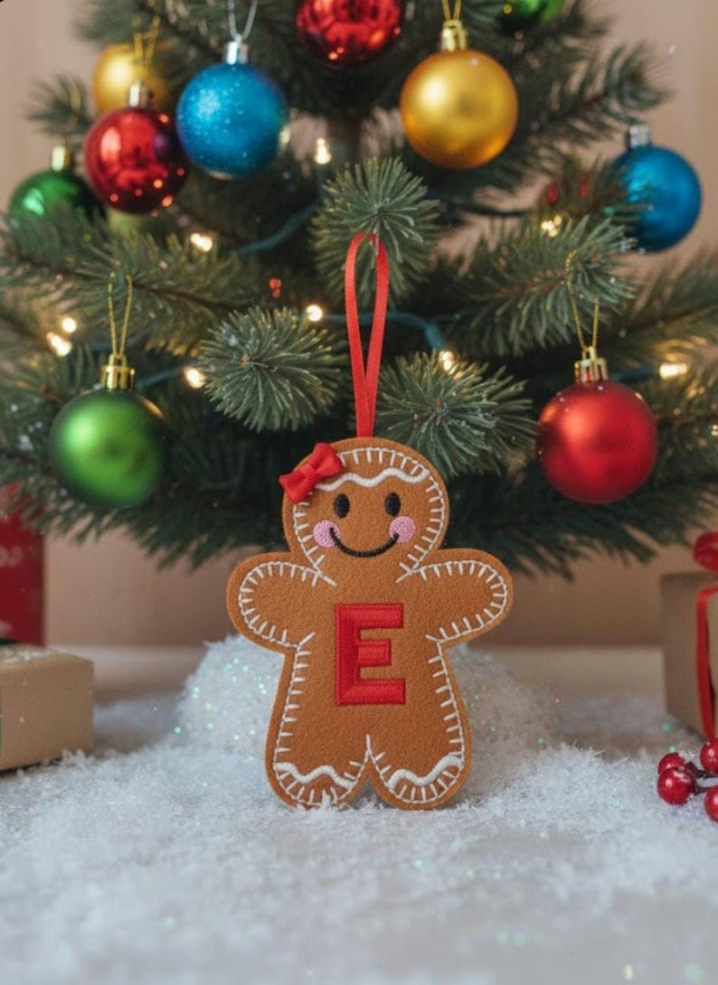May include: A brown felt gingerbread person ornament featuring a red letter E on the chest, a small red bow on the head, pink cheeks, and white embroidered icing details. The ornament hangs from a red ribbon loop against a Christmas tree.