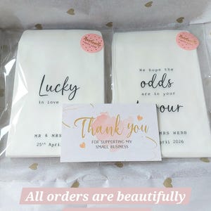 May include: Two white rectangular paper packets in clear plastic sleeves, each with a pink 'Handmade with love' sticker. The left packet reads 'Lucky in love MR & MRS 25th April'. The right reads 'We hope the odds are in your favour MRS WEBB April 2026'. A 'Thank you' card is centered in front.